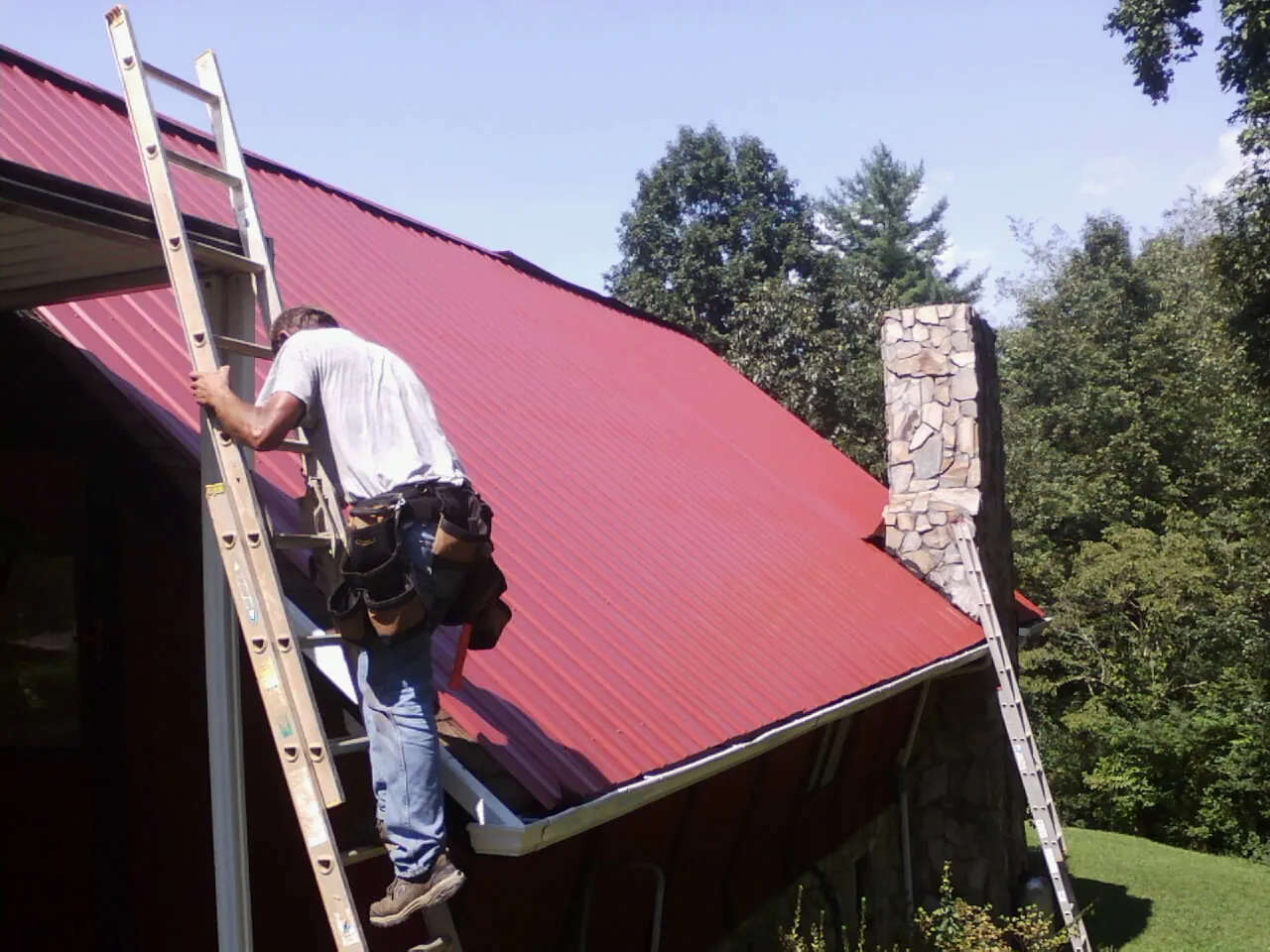 Roofer on a ladder beside a red metal roof and stone chimney — Soffit & Fascia Repair by Roofing Brothers in Derby